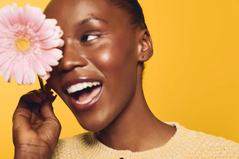 Smiling African American woman with dark skin holds pink flower near face against yellow background. Concept of happiness, beauty, natural skin, and floral wellness in studio portrait.