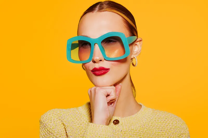 Stylish woman with oversized blue sunglasses and red lipstick poses confidently against bright yellow background. Fashion concept with bold colors and modern accessories.
