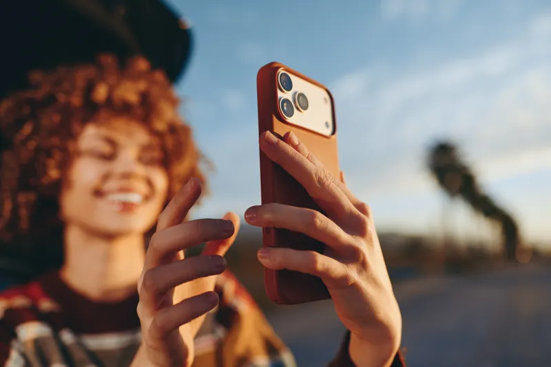 Woman lifestyle moment outdoors, holding a smartphone for a selfie, laughing with warm smile, curly hair blowing slightly, sunny street background with soft blur, candid travel vibe