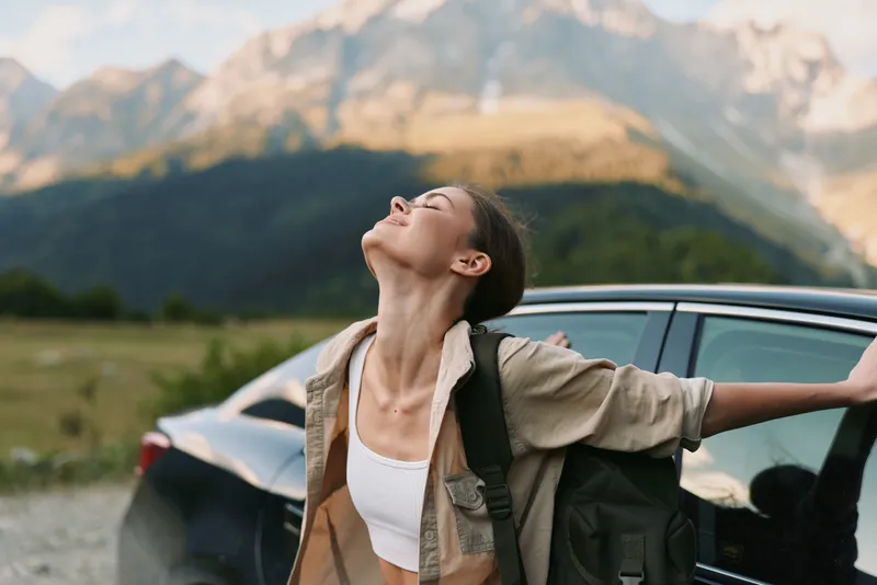 Joyful woman outdoors, leaning against a car with outstretched arms, sun in mountains background, lifestyle moment full of freedom and fresh air