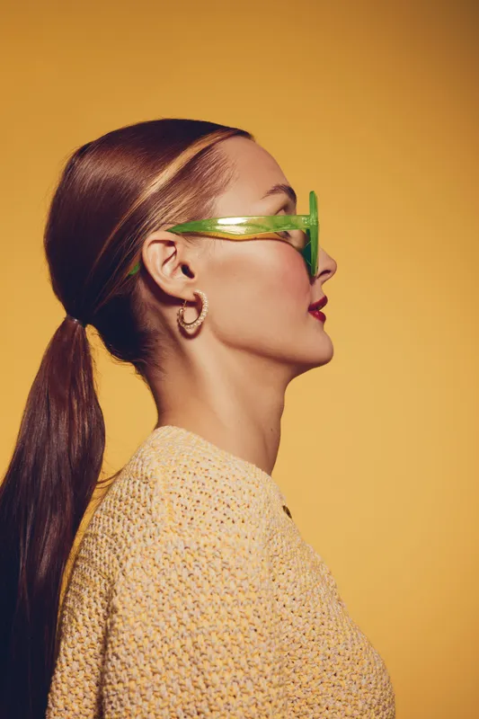 Profile of a stylish woman with green sunglasses and hoop earrings wearing a knitted sweater against a yellow background. Concept of fashion, accessories, and modern style.