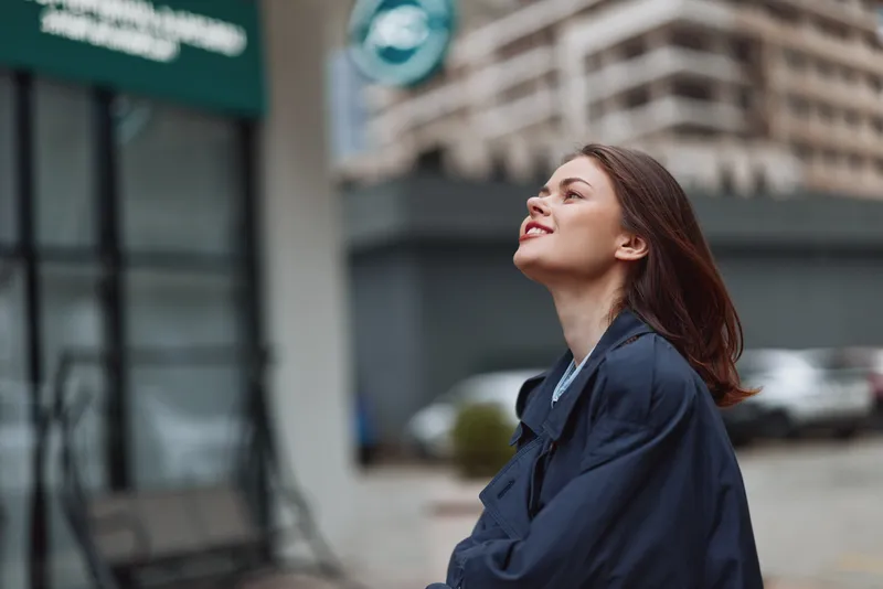 A young woman in a dark coat looks up smiling on a city street with blurred buildings in the background during daytime.