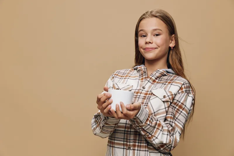 Girl in plaid shirt holding white bowl and spoon with smile against beige background. Child standing with relaxed posture and enjoying warm beverage or snack indoors.