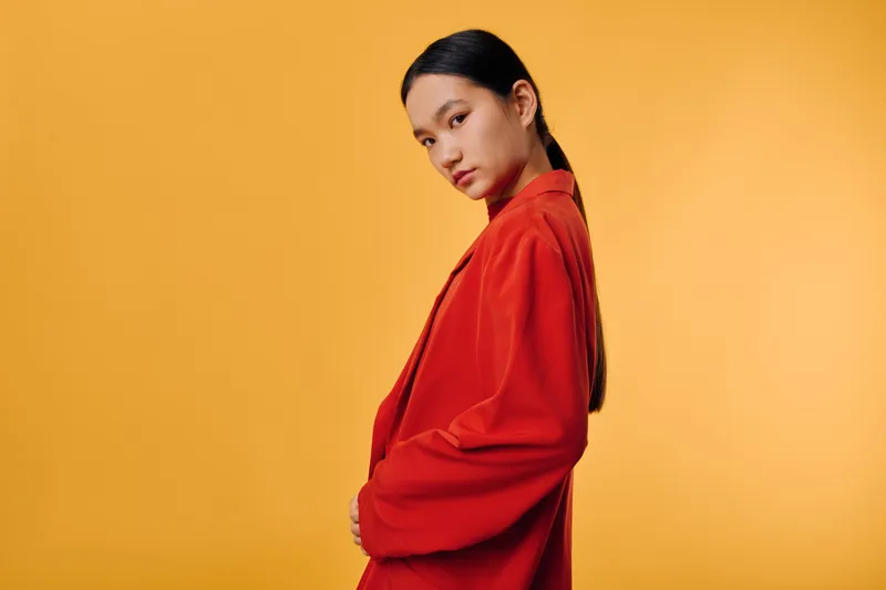 Woman with long dark hair wearing red blazer poses sideways on solid orange background for fashion, portrait, style, and beauty concept in studio lighting and simple setting.
