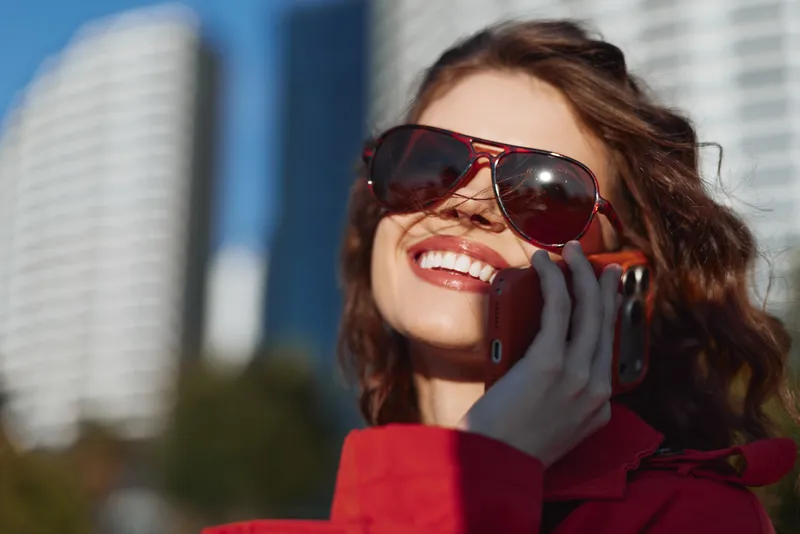 A smiling woman in a red jacket talks on a cellphone on a sunny urban street, with tall buildings behind her and a sense of casual daily connection.