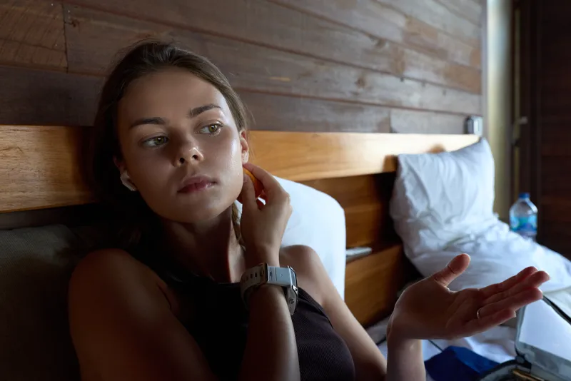 A tired woman sits on a bed in a wooden room, hand at her temple and the other gesturing as if in conversation. A moment of stress, fatigue, and reflection in a casual home setting.
