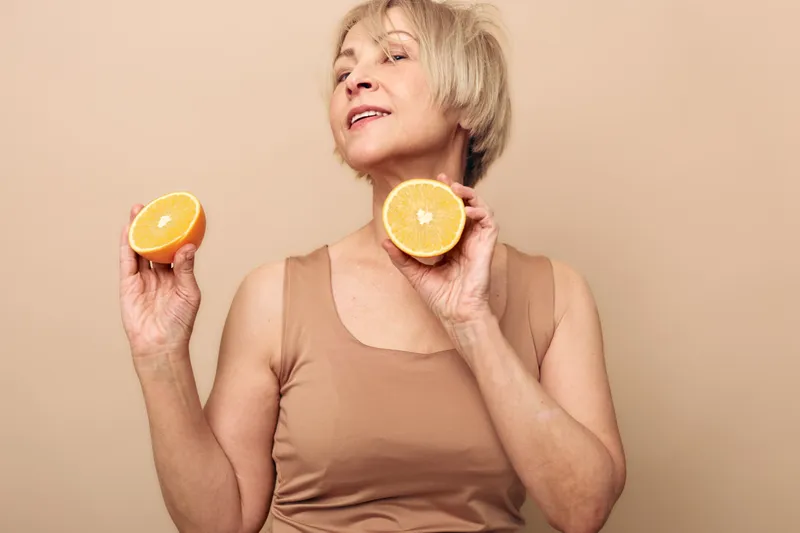 Mature woman in beige tank top holds two halves of fresh orange near face, posing confidently on neutral background. Concept for healthy lifestyle, vitamin C, and natural beauty.