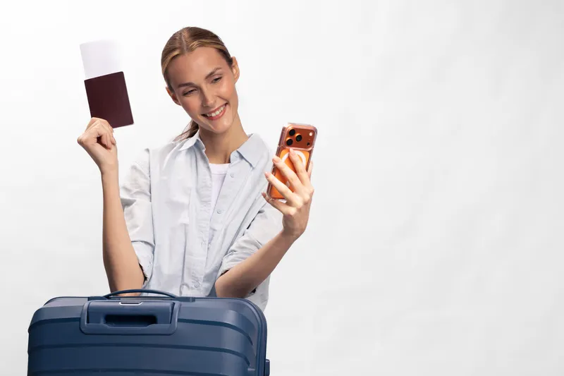 Smiling woman with long hair holds passport and phone near blue suitcase, preparing for travel. White background highlights her casual outfit and positive mood before trip.