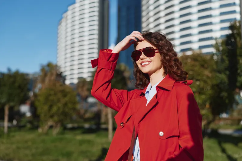Young woman in a red trench coat shields her eyes with sunglasses in a sunny park with modern high rise buildings behind.