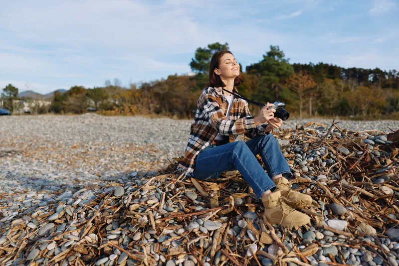 Woman sitting on a rocky shoreline, holding a camera, dressed in plaid shirt and boots, enjoying autumn light during a calm outdoor photography session.