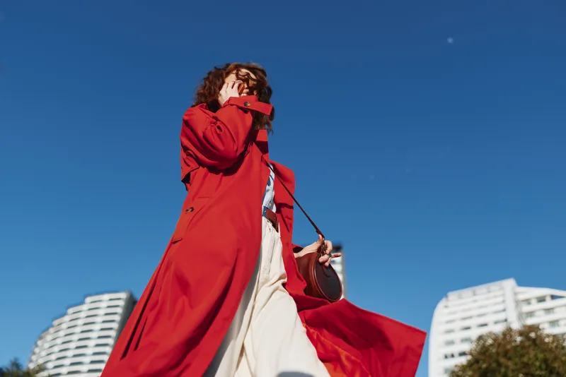 Young woman with curly hair wearing a flowing red coat walks confidently outdoors under a clear blue sky in an urban environment.