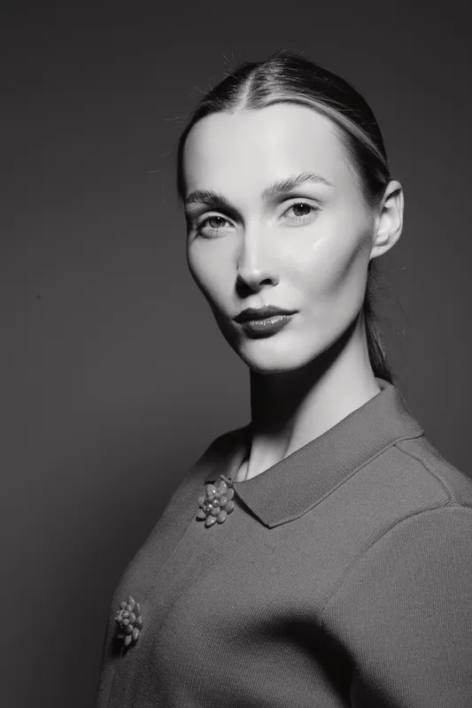 Portrait of a Caucasian woman with fair skin and slicked back hair wearing a textured top, looking confidently at the camera in studio lighting.