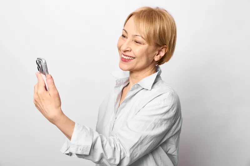 Woman senior smartphone smiling communication technology portrait device, elderly woman in light shirt holds a smartphone and smiles on white background, studio indoor lifestyle