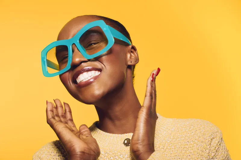 Happy African woman with dark skin wearing large blue sunglasses and yellow sweater smiles with closed eyes on bright yellow background. Concept of joy, fashion, and summer style.