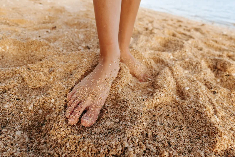 Close up of bare feet partially buried in dry sand on a sunny beach near calm water. Concept for summer, relaxation, vacation, and natural outdoor environment.