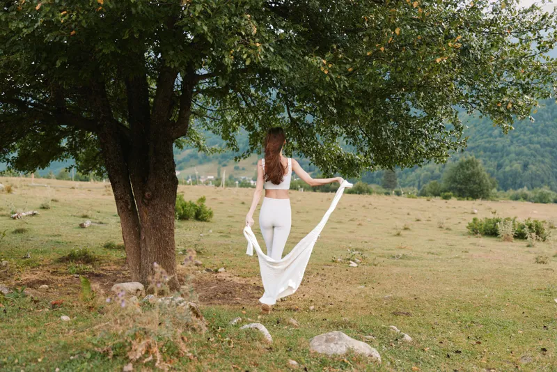 A woman in white activewear stands barefoot on grass near a tree holding a flowing white fabric in a natural outdoor setting with mountains in the background.