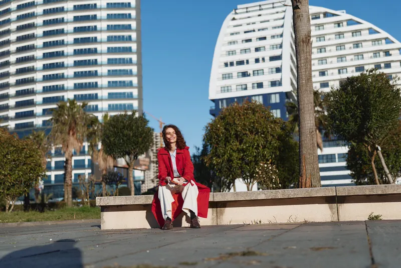 Smiling woman sitting outdoor city fashion red coat, candid pose on a low wall with modern buildings behind, warm daylight and casual boots create a cheerful mood