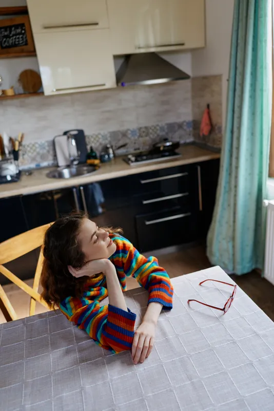 A woman sits at a kitchen table with her chin resting on her hand, wearing a colorful striped sweater, while natural light fills the room, creating a warm, contemplative mood and a cozy home moment.