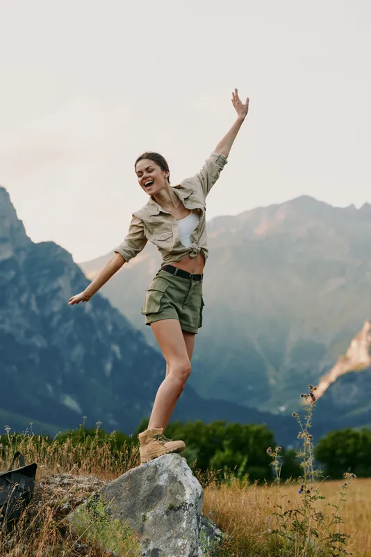 Cheerful woman outdoors, jumping on a rock, carefree moment, sunny day, mountains in background, casual outfit, natural colors, energetic pose, sense of adventure