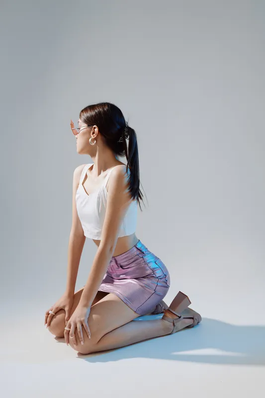 Young woman in fashionable outfit kneels on the floor looking sideways. Studio shot with soft lighting highlights beauty, fashion, and style concepts.