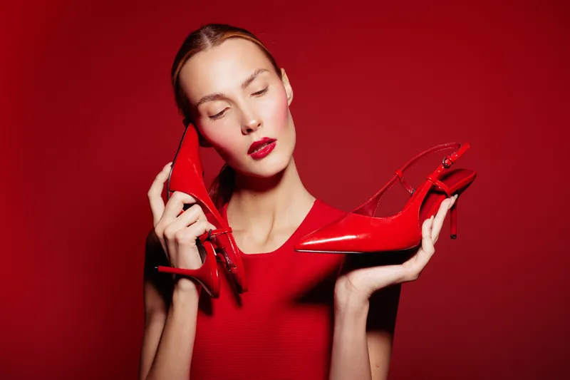 Young woman in red dress holds red high heels near face with closed eyes on red background. Concept of fashion, style, elegance, and beauty in studio lighting.