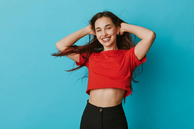 Young woman with long hair wearing red crop top and black pants posing with hands behind head smiling confidently against blue background in studio portrait.