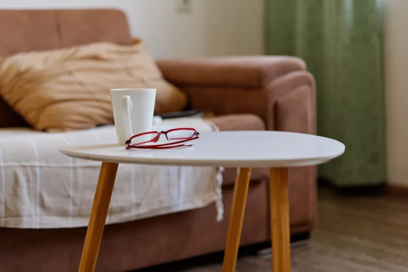 Cozy living room scene with a white round table, mug, red glasses, soft sofa in the background, natural light creating a warm relaxed home atmosphere.
