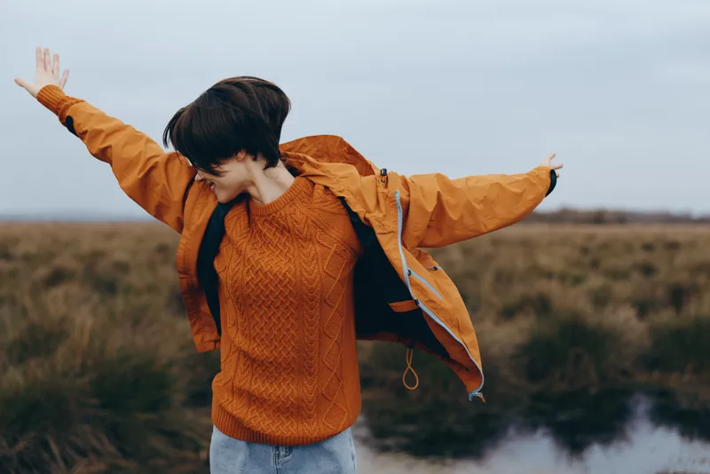 Child in autumn outdoors, arms spread, wearing coat and jacket, by lake landscape with windy nature, warm orange color, joy lifestyle in cloudy sky fresh near river countryside leisure.