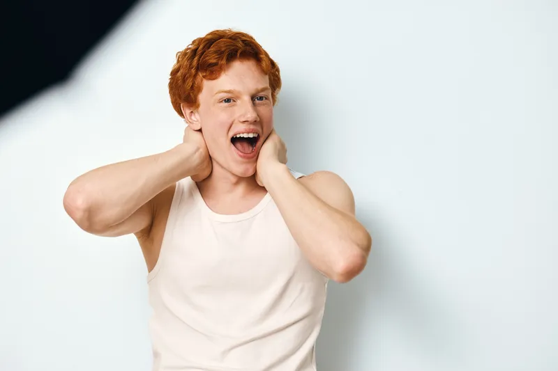 Young man with red hair in white tank top standing with hands on neck expressing surprise and excitement against light background in studio.