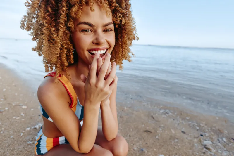 Woman on beach, curly hair, smile and laugh; summer sea leisure, happiness, closeup portrait, warm sunlight, calm waves behind.