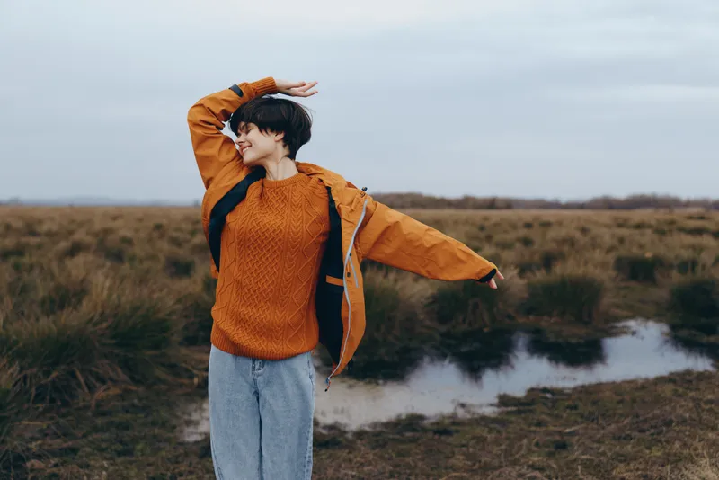 Woman lifestyle outdoor portrait in autumn, orange sweater, standing near river bank, smiling and stretching sleeves, cloudy sky, soft light, natural field background, casual pose, by water