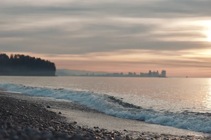 Beach at dusk with gentle waves lapping the pebbled shore, distant city skyline on the horizon, warm tones reflecting on calm water and cloudy sky.