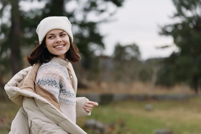 Woman in a cozy knit sweater and light coat walking outdoors with a white beanie, smiling as she enjoys a chilly day in nature with trees and open field