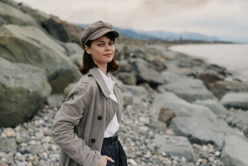 Young woman in stylish checkered coat and cap stands on rocky beach with calm sea and cloudy sky in background. Concept of fashion, autumn, and outdoor lifestyle portrait.