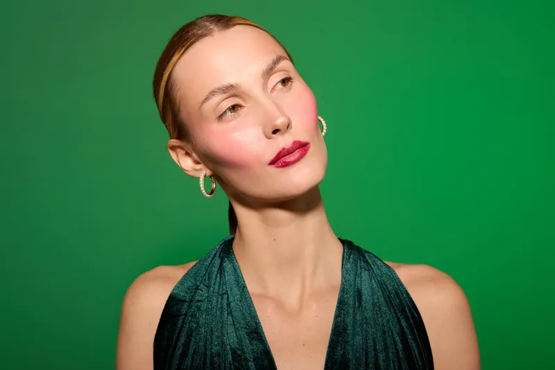 Portrait of elegant woman with red lipstick and hoop earrings wearing dark green dress on green background. Concept of beauty, fashion, and confident female model.