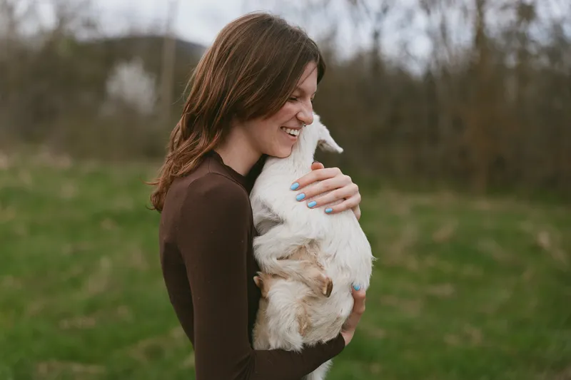 Goat, kid, woman, outdoor, lifestyle, affection, animal, field, rural, hug appear as a smiling woman embraces a white goat kid in a grassy open area, conveying warm connection and gentle countryside