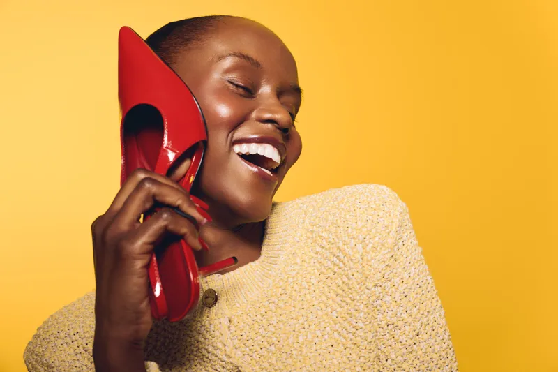 Smiling dark skinned African woman in beige sweater holds red high heel shoe near face on yellow background. Concept for fashion, beauty, joy, and lifestyle with vibrant colors.