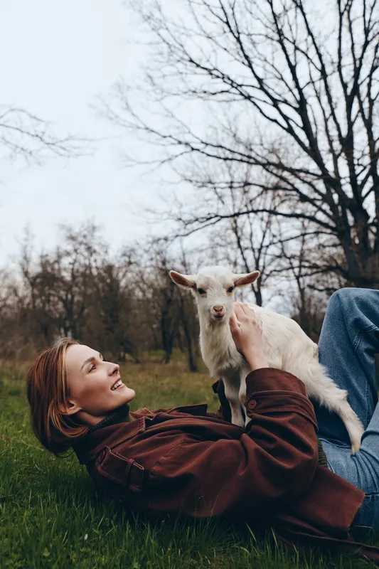 Relaxed woman lies on grass in a park, stroking a playful goat kid, enjoying outdoor life and gentle connection with nature, candid moment of simple countryside joy