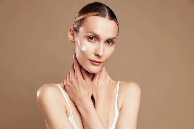 Young woman applies facial cream on clean skin with hands touching neck, looking at camera, beauty skincare routine for healthy glowing complexion isolated on beige background.