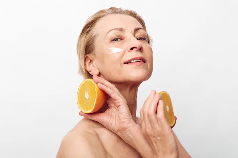Mature woman with natural skin care cream applied under eyes holds two orange halves near her face, smiling softly on white background. Concept of healthy skin and natural beauty.