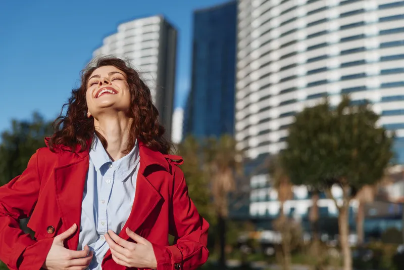 Young woman with curly hair enjoys sunny weather in the city wearing a red coat and light blue shirt with a joyful expression and closed eyes.