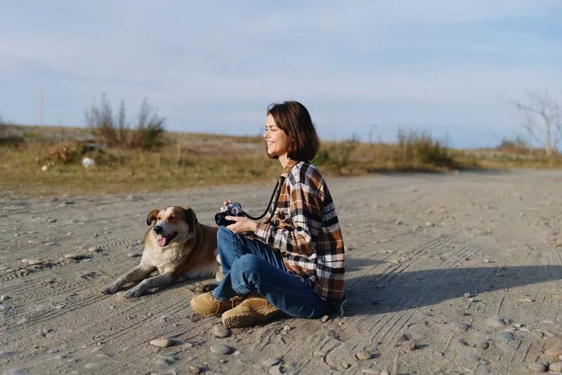 Woman sitting on a sandy beach with a dog, holding a camera, enjoying outdoor photography in a calm seaside setting, plaid shirt, jeans, sunlit horizon and peaceful companionship.