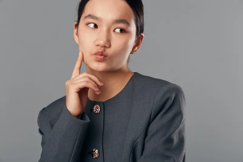 Young woman in gray jacket making thinking gesture with finger on cheek looking sideways with pursed lips and contemplative expression on plain gray background studio portrait.
