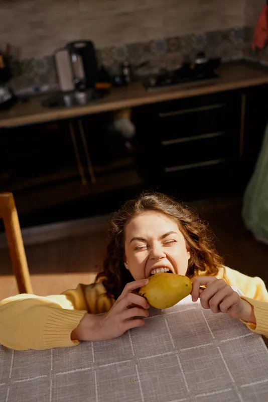 A woman in a yellow sweater bites into a mango over a checkered tablecloth in a cozy kitchen, conveying casual home life and playful indulgence