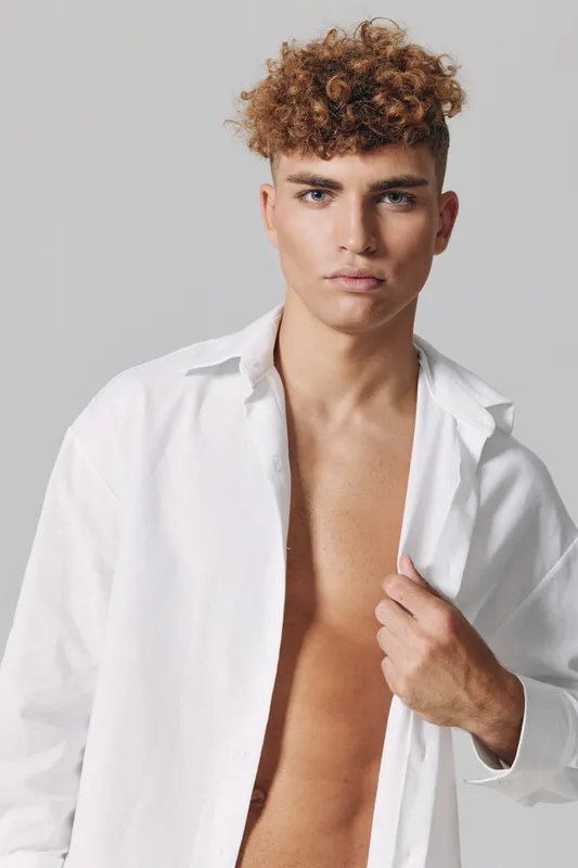 Portrait of young man with curly hair wearing open white shirt looking confident against plain gray background. Male model with brown skin in studio lighting and casual pose.