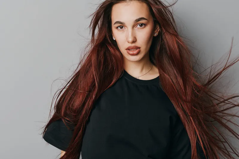 Young woman with long red hair wearing black t shirt standing against gray background, looking at camera with confident expression and natural makeup in studio portrait.