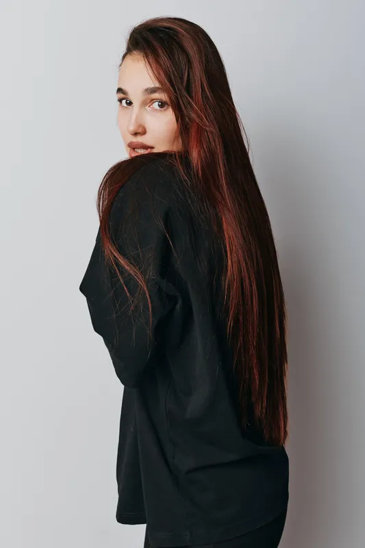 Young woman with long brown hair wearing black sweater standing against white background looking over shoulder with calm expression and natural makeup in studio portrait.