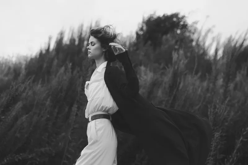 Wind sweeps as a cape trails behind a poised woman in a flowing dress across a grassy field, capturing dramatic movement, outdoors silhouette, and monochrome elegance in an atmospheric scene