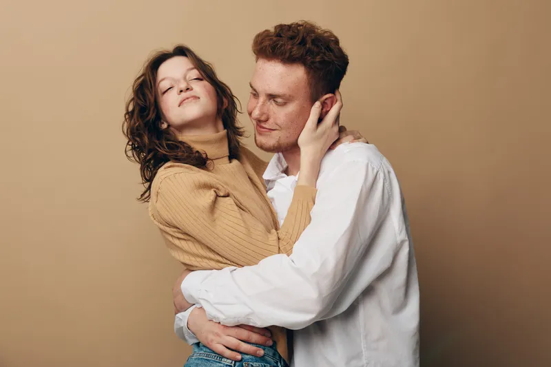 Young couple embraces tenderly against beige background. Woman in brown sweater closes eyes while man in white shirt holds her gently. Concept of love, affection, intimacy, and connection.