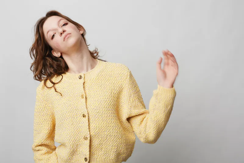 Young woman in yellow knitted cardigan poses with raised hand and relaxed expression against plain grey background. Casual fashion, portrait, lifestyle, and natural beauty concept.